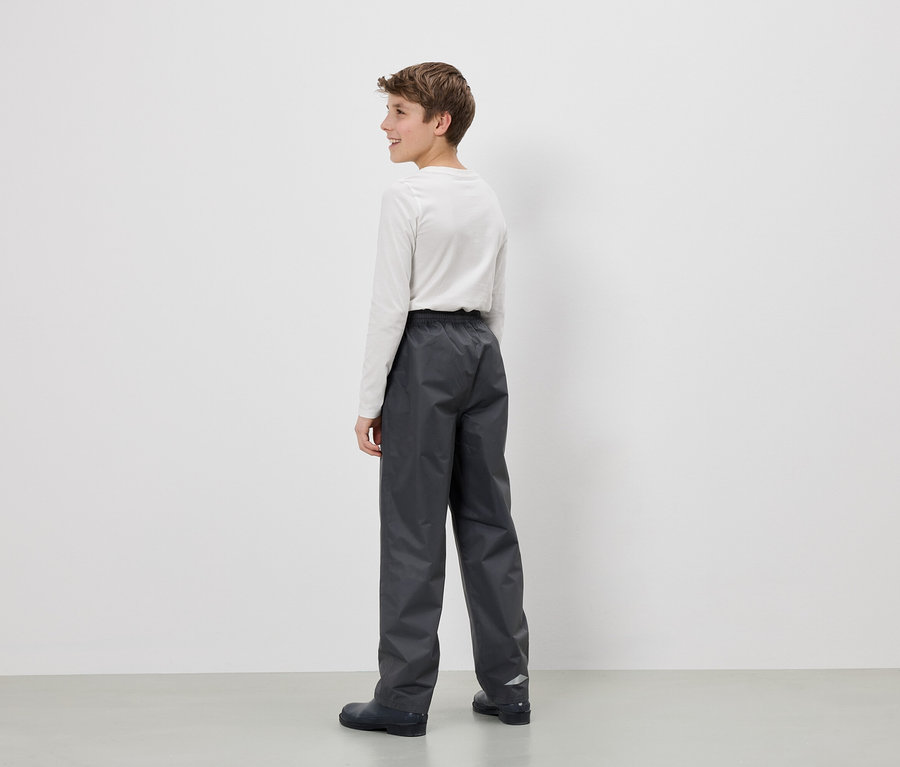 Un adolescent se tient devant un mur blanc, portant un pantalon de pluie gris et un t-shirt blanc à manches longues.