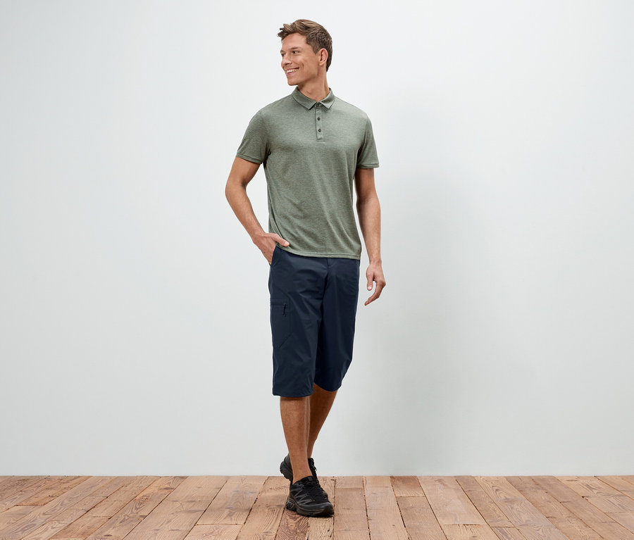 Un homme se tient debout, une main dans la poche, devant un mur blanc, portant un polo vert, un short bleu et des chaussures noires.