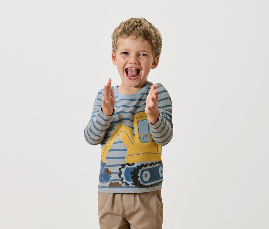 Un garçon blond applaudit, portant un t-shirt à manches longues bleu rayé avec un bulldozer jaune.
