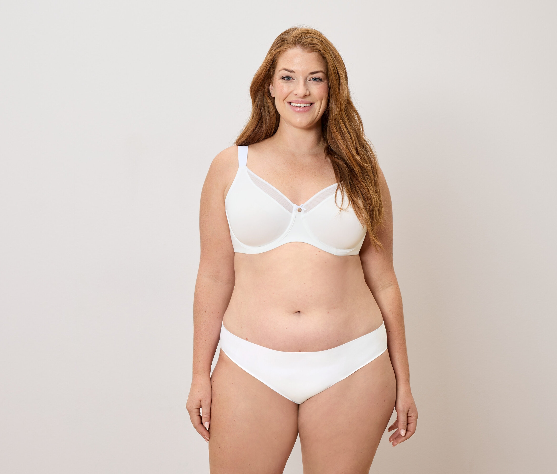 Femme aux longs cheveux roux portant un soutien-gorge et une culotte blancs, souriant.