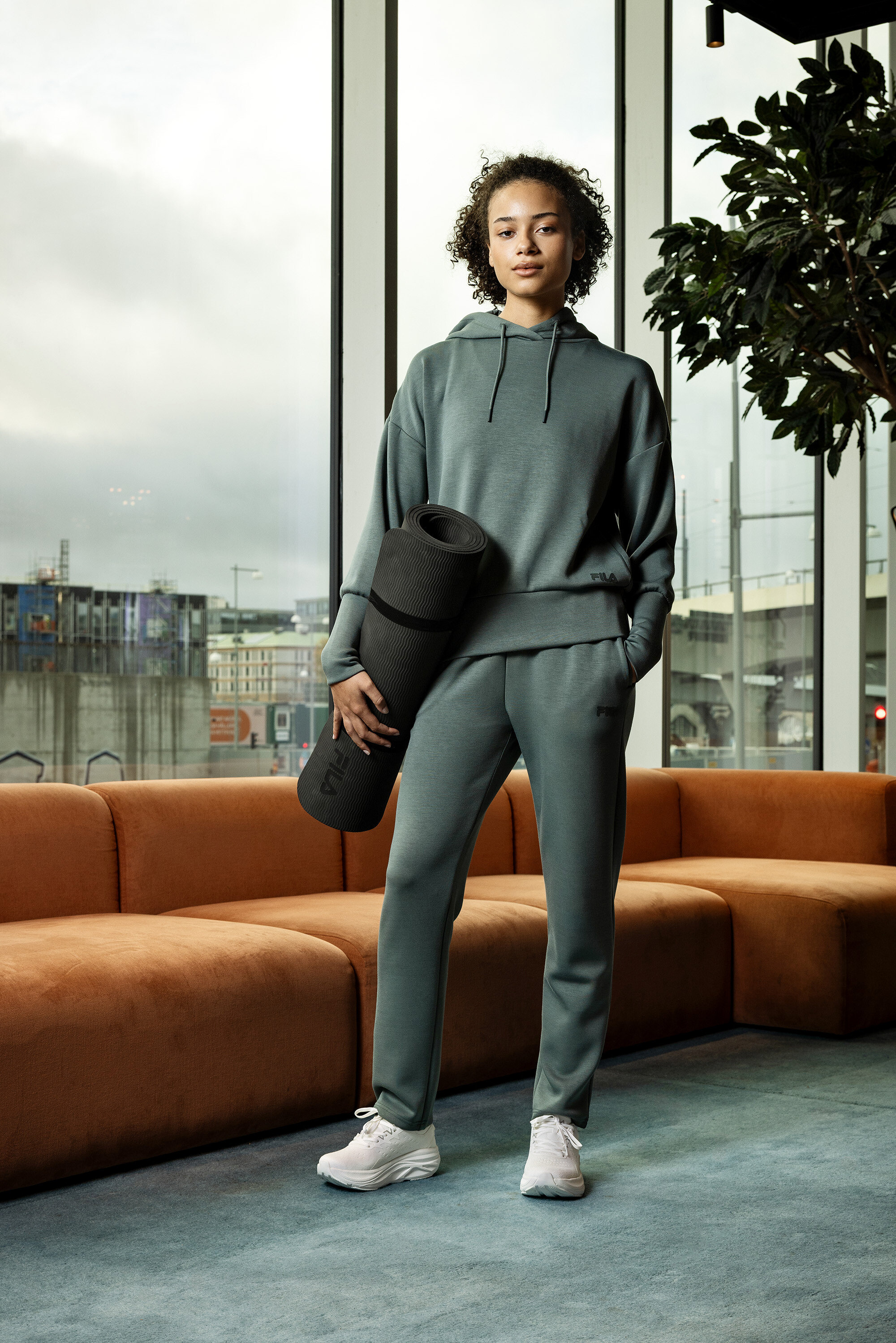 Jeune femme en survêtement gris tenant un tapis de yoga enroulé devant une fenêtre.