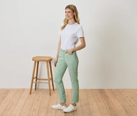 Une femme se tient à côté d'un tabouret et porte un t-shirt blanc, un pantalon vert et des baskets blanches.