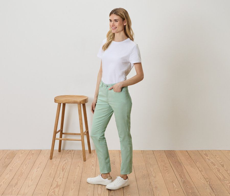 Une femme se tient à côté d'un tabouret et porte un t-shirt blanc, un pantalon vert et des baskets blanches.