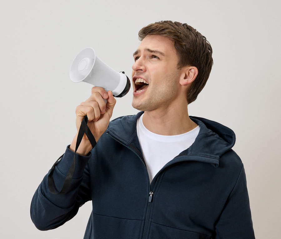 Un homme aux cheveux bruns crie dans un mégaphone blanc. Il porte un sweat à capuche bleu marine et un t-shirt blanc.