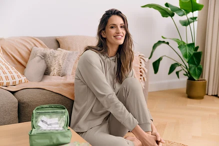 Femme assise sur un tapis tressé dans le salon, portant un survêtement beige, souriant. Plante, canapé, trousse de toilette verte à l'arrière-plan.