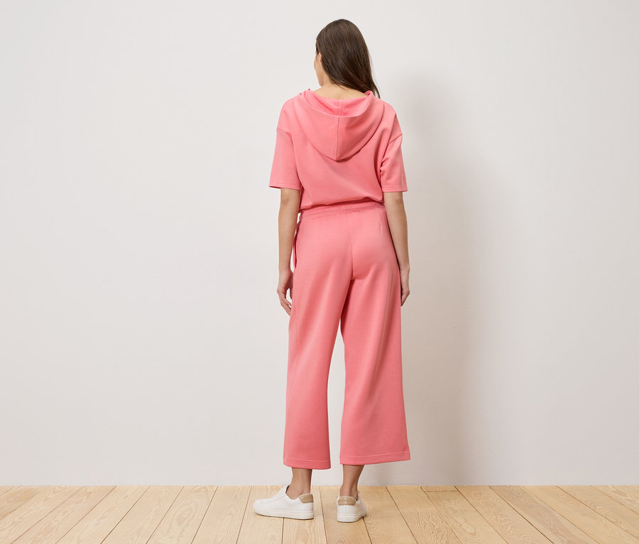 Une femme se tient dos à la caméra, portant un sweat à capuche rose et un pantalon culotte.