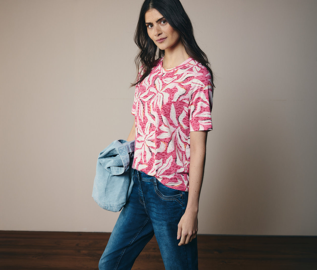 Une femme porte un t-shirt à manches courtes CECIL rose à motifs et un jean. Une veste en jean est suspendue à son épaule.