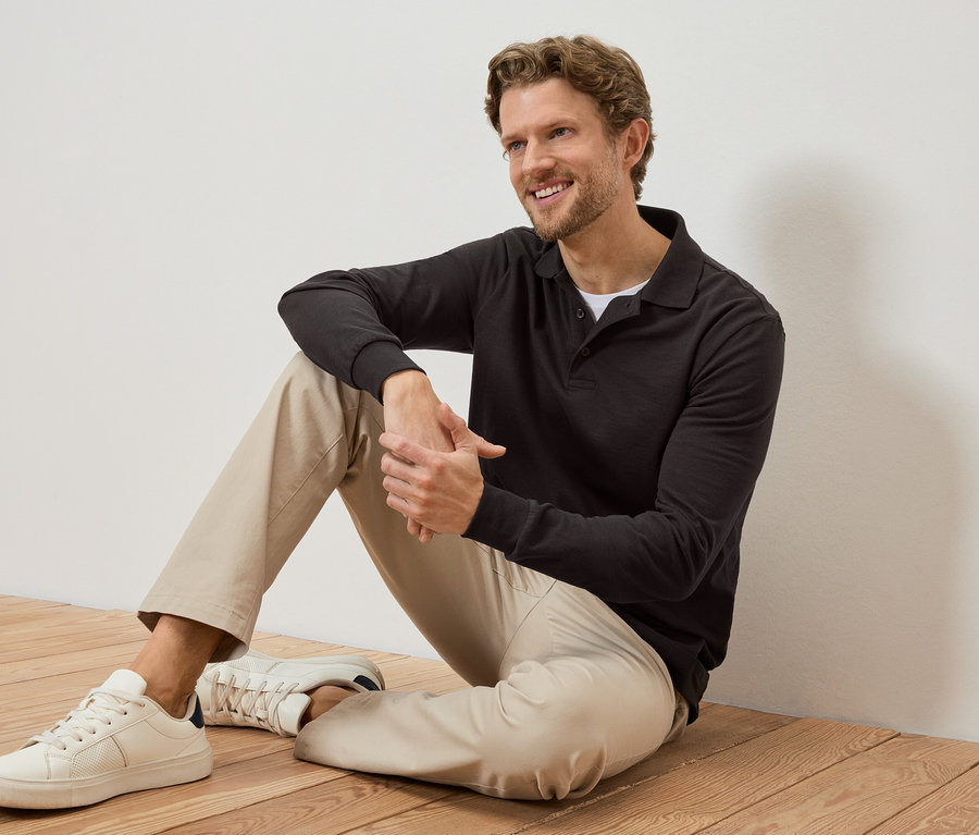 Un homme souriant est assis sur un plancher en bois, portant un polo foncé, un pantalon clair et des baskets blanches.