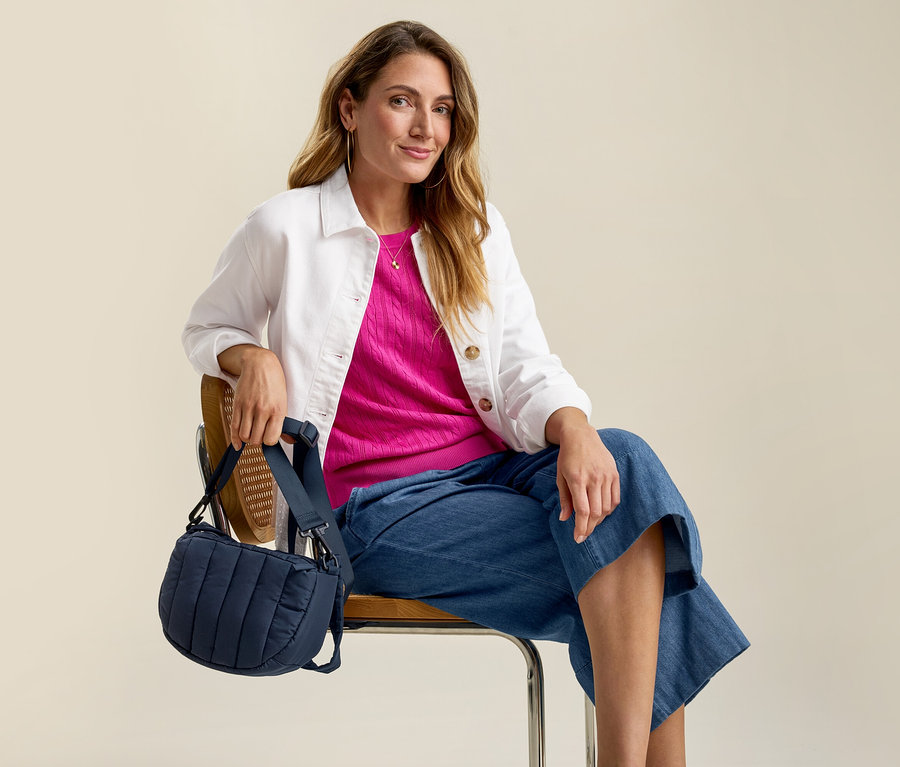 Femme assise sur une chaise avec veste en jean avec broderie ajourée, haut rose, jean et sac bleu.