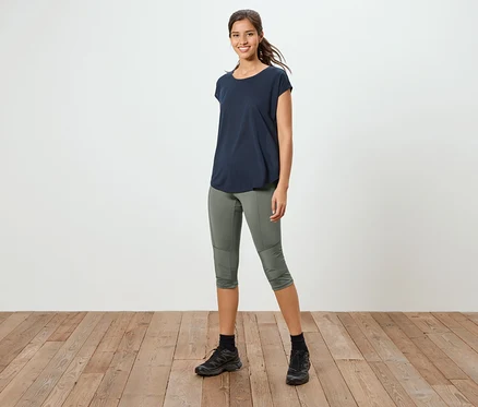 Une femme se tient sur un plancher en bois, portant un t-shirt bleu marine, un pantalon 3/4 vert olive et des baskets noires.
