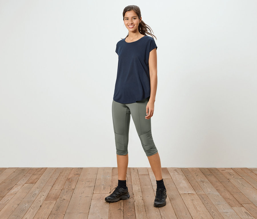 Une femme se tient sur un plancher en bois, portant un t-shirt bleu marine, un pantalon 3/4 vert olive et des baskets noires.