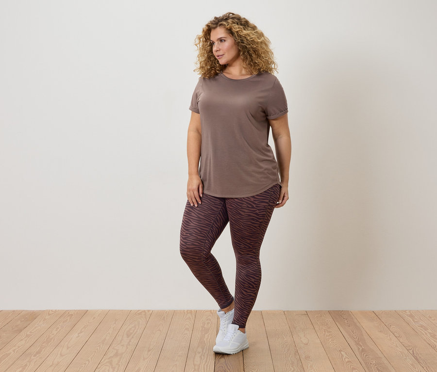 Femme aux cheveux bouclés portant un t-shirt marron et des leggings à motifs posant devant un mur.