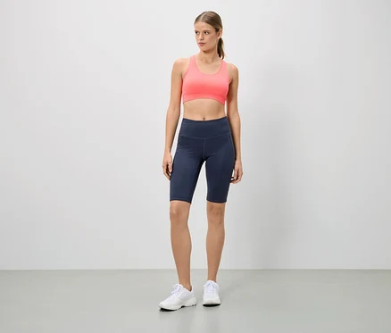 Une femme debout porte une tenue de sport composée d'un soutien-gorge de sport rose, d'un cycliste bleu marine et de baskets blanches.