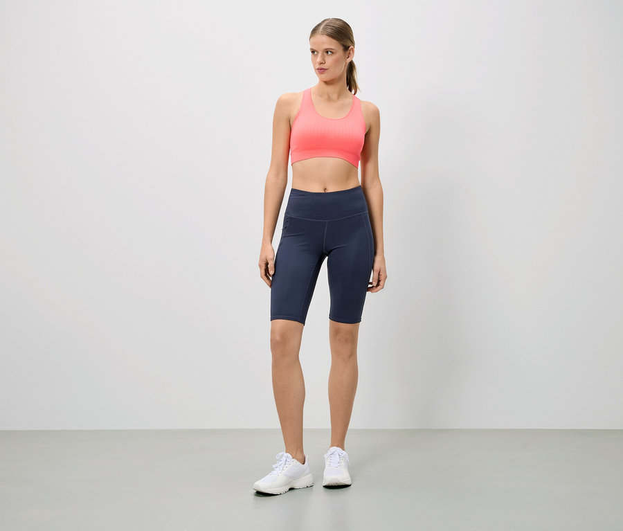 Une femme debout porte une tenue de sport composée d'un soutien-gorge de sport rose, d'un cycliste bleu marine et de baskets blanches.