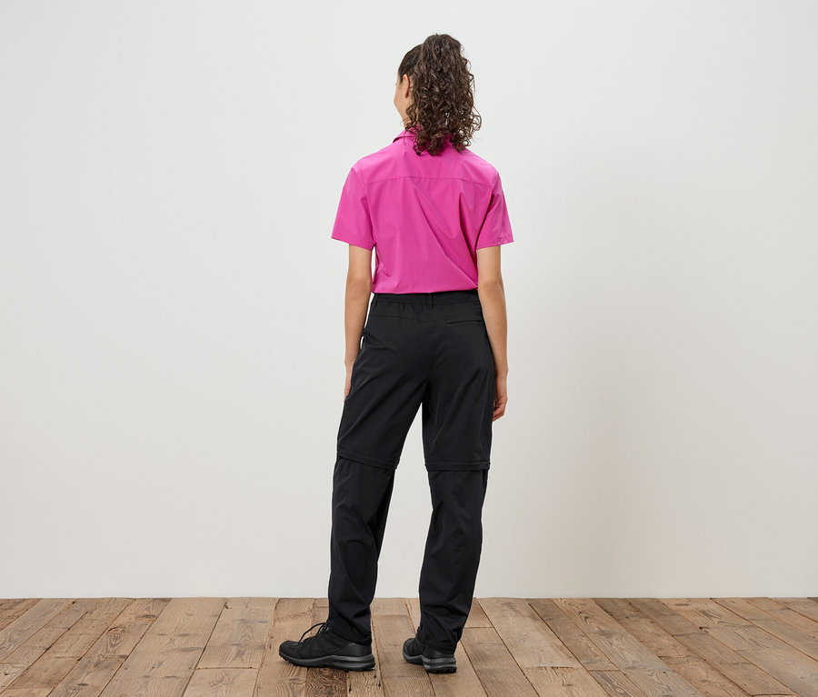 Vue arrière d'une femme aux cheveux bouclés portant une chemise rose et un pantalon noir zippé, avec des chaussures noires.