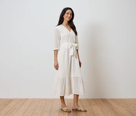 Une femme se tient devant un mur blanc et porte une robe blanche avec une ceinture et des sandales.