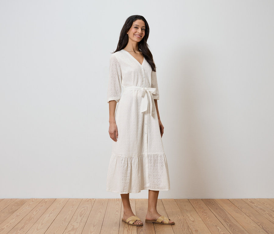 Une femme se tient devant un mur blanc et porte une robe blanche avec une ceinture et des sandales.