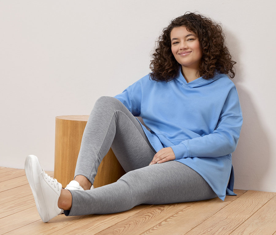 Une femme aux cheveux bouclés est assise par terre, portant un sweat à capuche bleu clair, un legging gris et des baskets blanches.