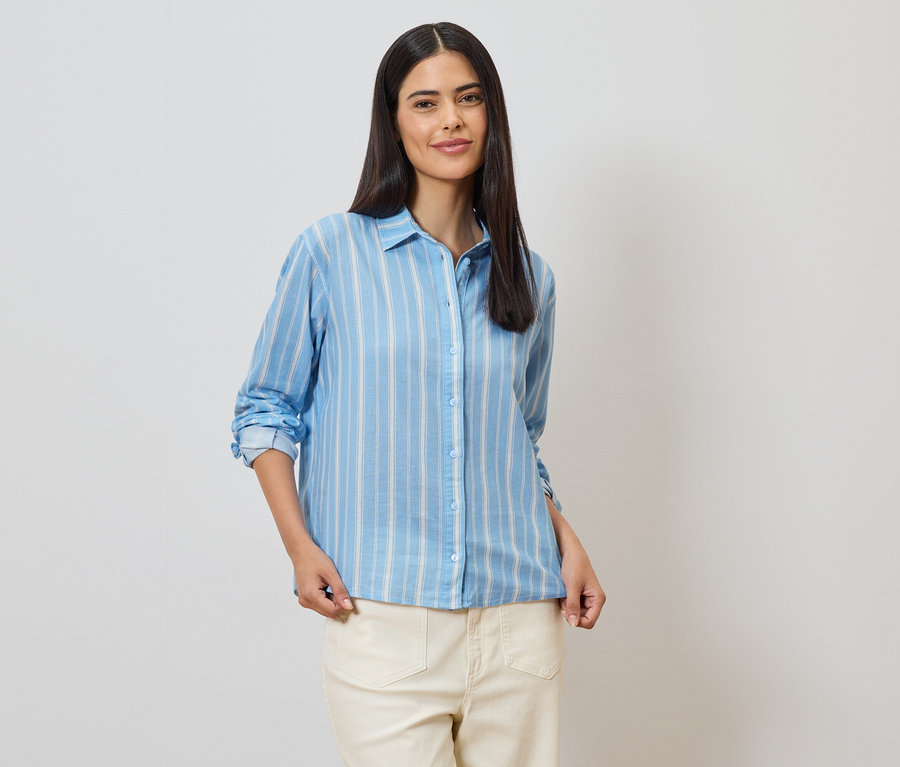 Une femme aux longs cheveux foncés porte une chemise rayée bleu clair et un pantalon clair sur un fond blanc.