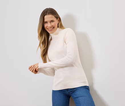 Une femme blonde souriante porte un pull à col roulé crème et un jean bleu devant un mur blanc.