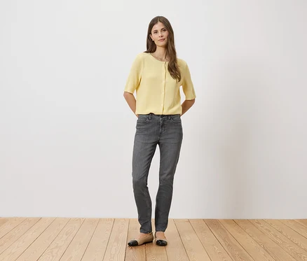 Une femme en chemisier jaune et jean gris, debout devant un mur blanc sur un plancher en bois.