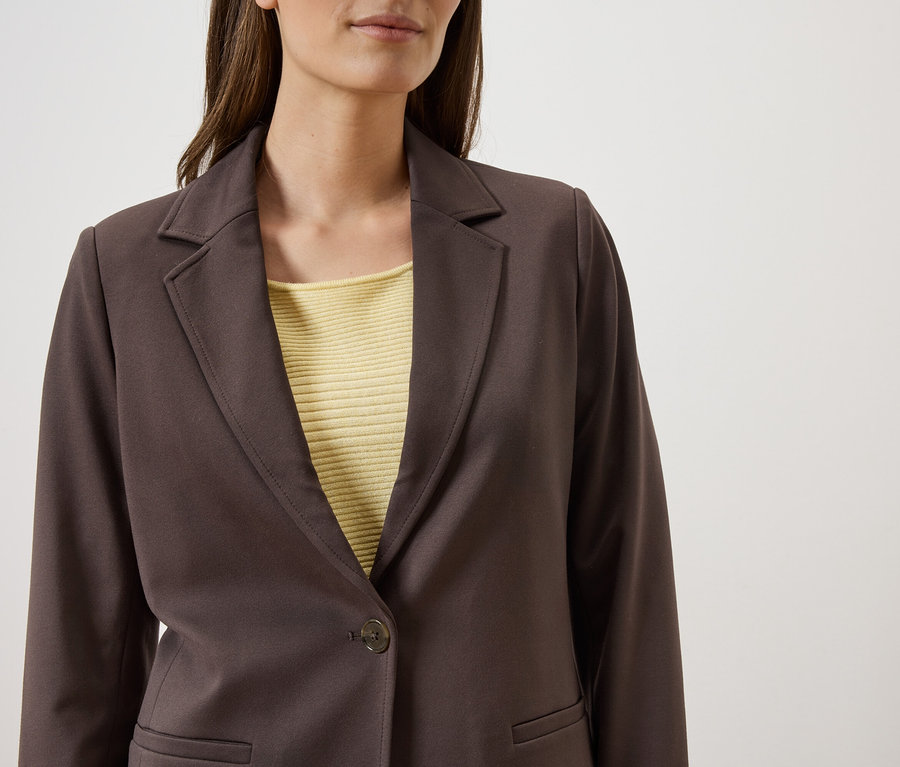 Une femme porte un blazer marron sur un haut jaune devant un fond blanc.