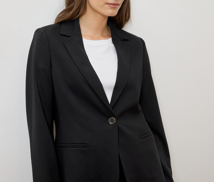Une femme porte un blazer noir sur un t-shirt blanc sur fond blanc.