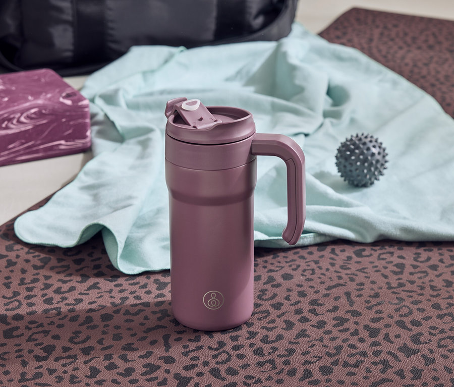 Une gourde isotherme violette avec une poignée est posée sur un tapis de yoga marron à motif léopard. À côté se trouvent une balle de massage grise, une serviette turquoise et une brique de yoga violette.