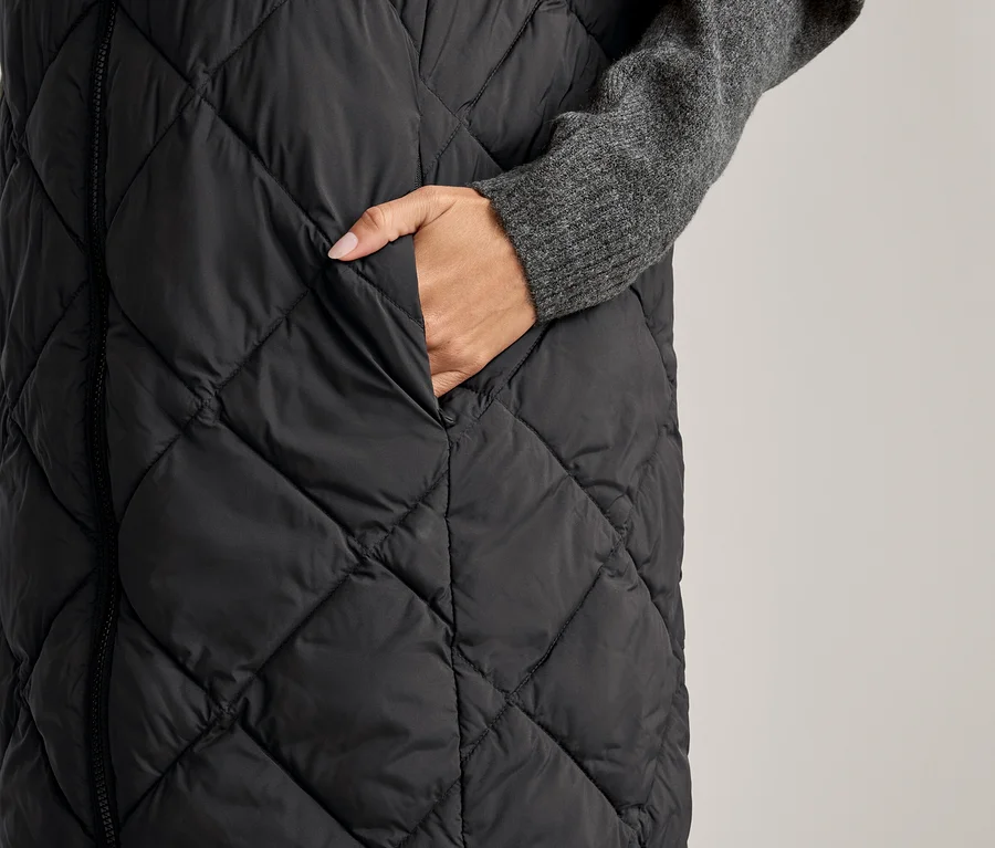 Une femme glisse sa main dans la poche d'un long gilet noir matelassé porté sur un pull gris.