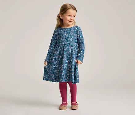 Une fillette blonde porte une robe bleue à fleurs à manches longues, des collants roses et des chaussures.