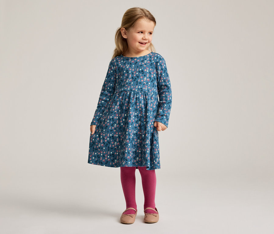 Une fillette blonde porte une robe bleue à fleurs à manches longues, des collants roses et des chaussures.