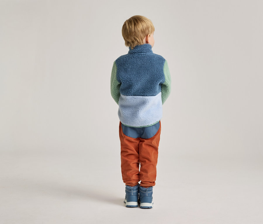 Vue arrière d'un enfant aux cheveux blonds courts portant une veste en polaire de couleur, un pantalon en velours côtelé orange et des bottes bleues.
