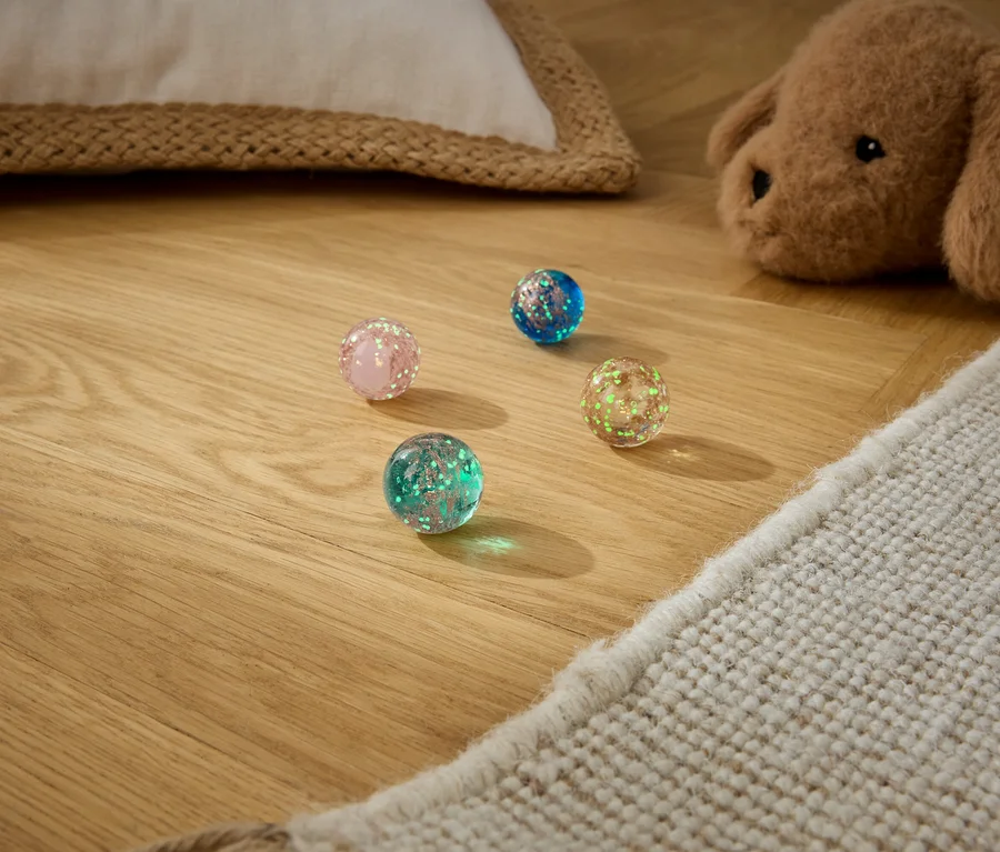 Quatre balles rebondissantes pailletées roses, bleues, vertes et jaune-vert sont posées sur un parquet en bois, à côté d'un tapis beige et d'un chien en peluche marron.