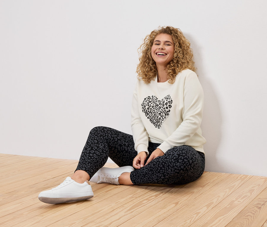 Une femme aux cheveux bouclés est assise sur un plancher en bois, portant un pull crème avec un cœur léopard noir, des leggings léopard et des baskets blanches.