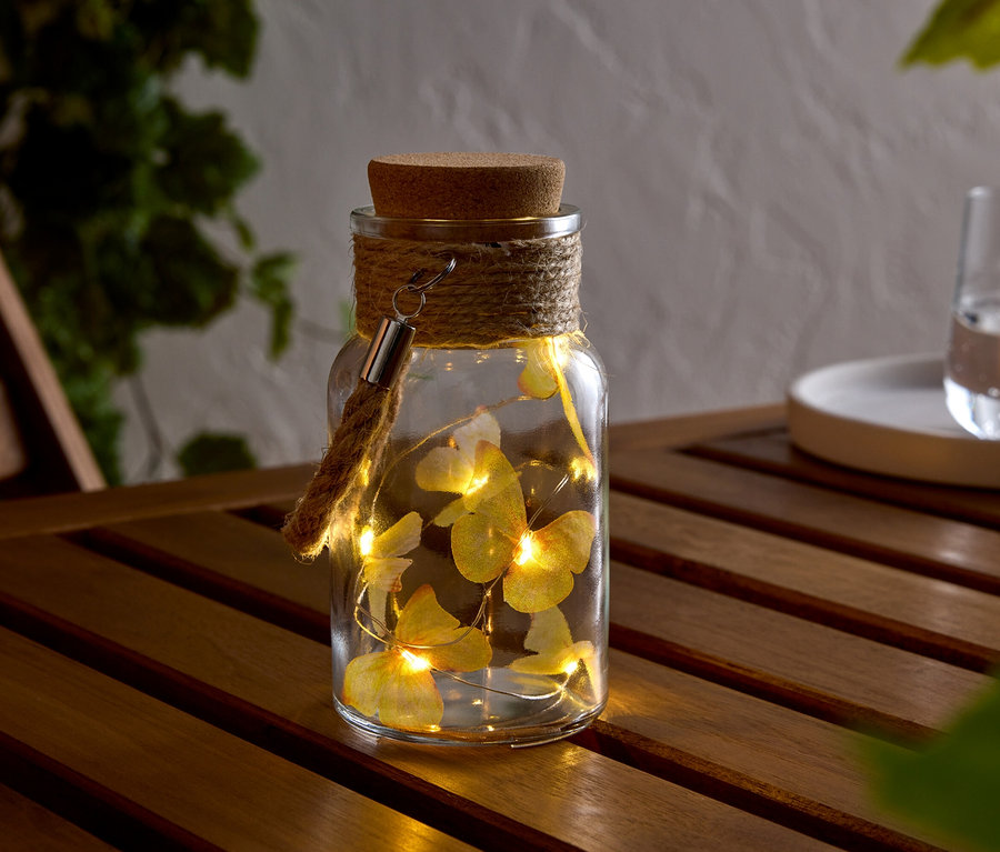 Bocal en verre avec bouchon en liège, entouré d'une corde, avec une guirlande lumineuse avec des papillons à l'intérieur, sur une table en bois.