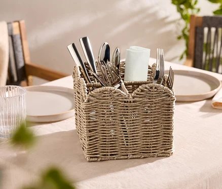 Un panier à couverts tressé contenant des couverts argentés et une serviette en tissu blanche est posé sur une table.