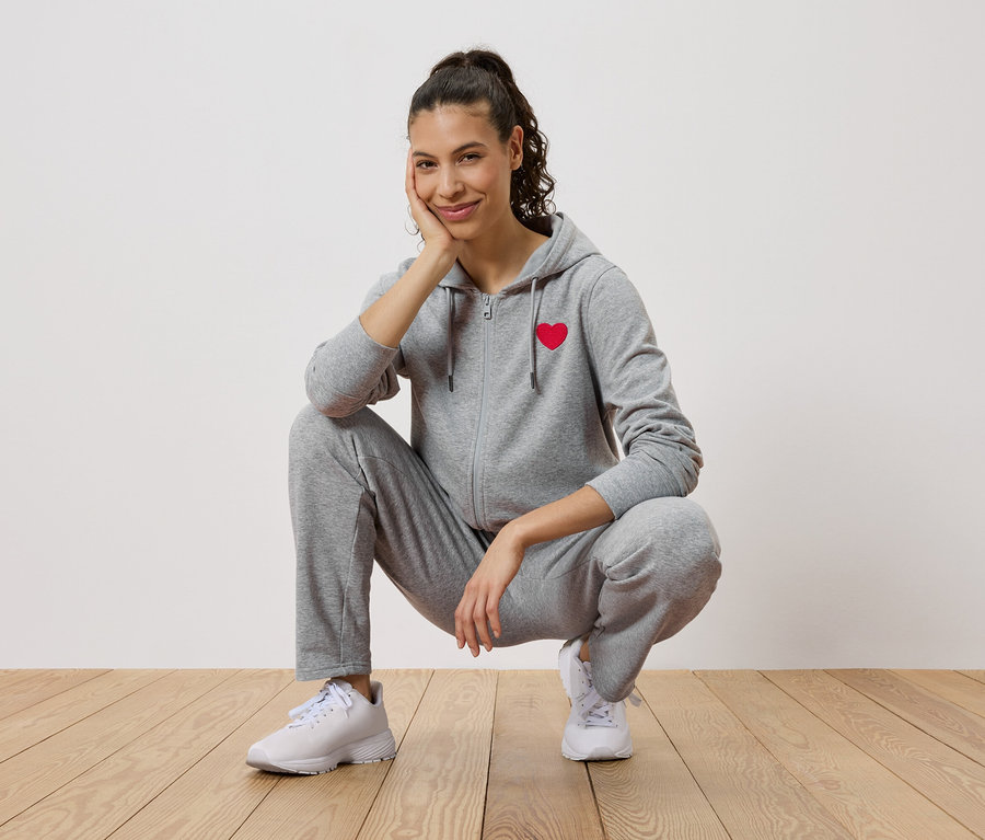 Femme accroupie sur un plancher en bois, portant un survêtement gris avec un cœur rouge et des baskets blanches.