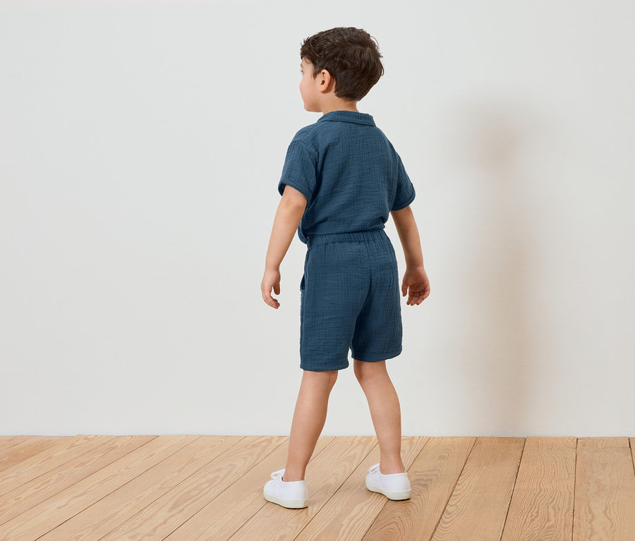 Garçon vu de dos portant un ensemble short en mousseline bleu et des chaussures blanches, sur un sol en bois devant un mur blanc.