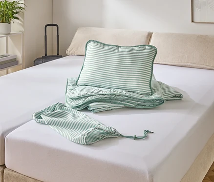 Un lit avec un drap-housse blanc, sur lequel reposent un oreiller rayé, une couverture et un sac.