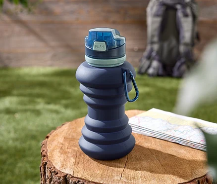 Une gourde bleue pliable avec un bouchon bleu est posée sur une souche d'arbre. Une carte de randonnée et un sac à dos sont visibles en arrière-plan.
