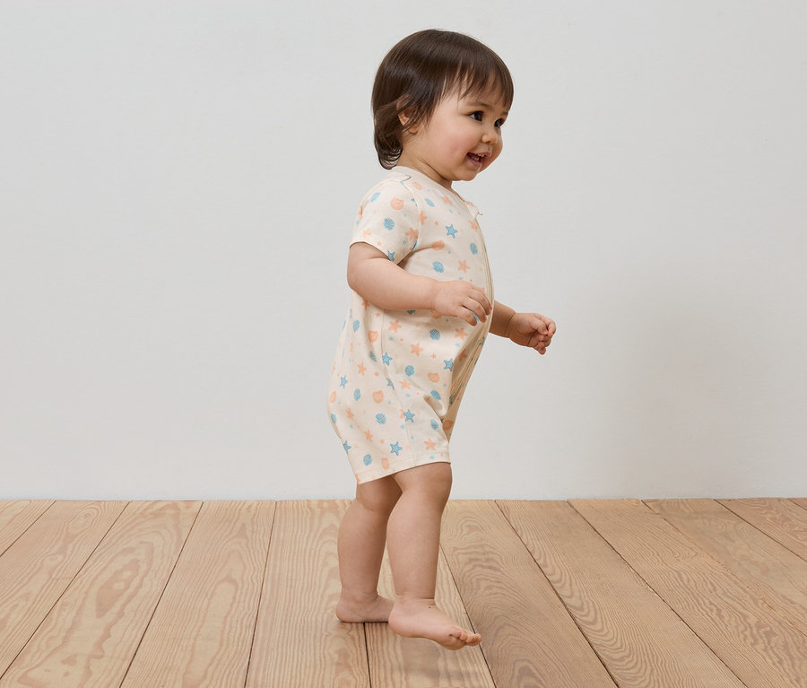 Un jeune enfant aux cheveux bruns porte une barboteuse claire à motifs bleus et oranges et marche sur un plancher en bois.
