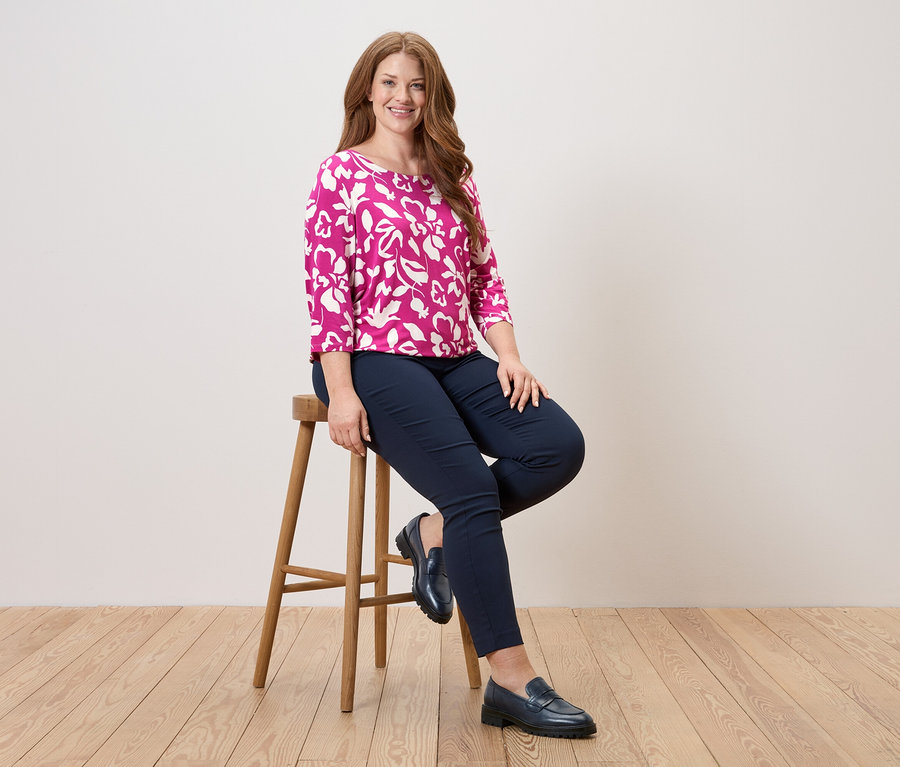Femme assise sur un tabouret, portant un chemisier rose à motifs floraux, un pantalon bleu et des chaussures en cuir.
