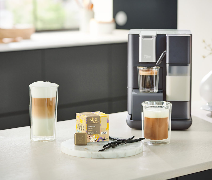 Un Qbo MILK MASTER Gentle Grey est placé à côté de deux verres de café. Un Qbo MILK MASTER rouge est visible en arrière-plan.