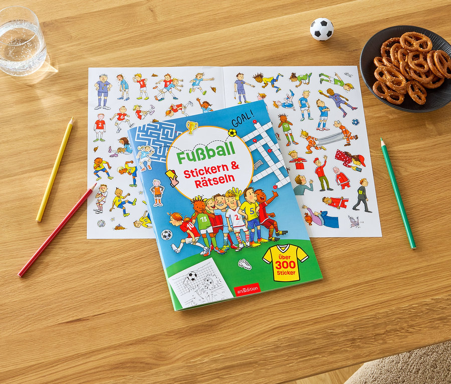 Sur une table en bois se trouvent un verre d'eau, un bol de bretzels, un ballon de football, des crayons de couleur et un livre de stickers et d'énigmes sur le football avec une planche de stickers.