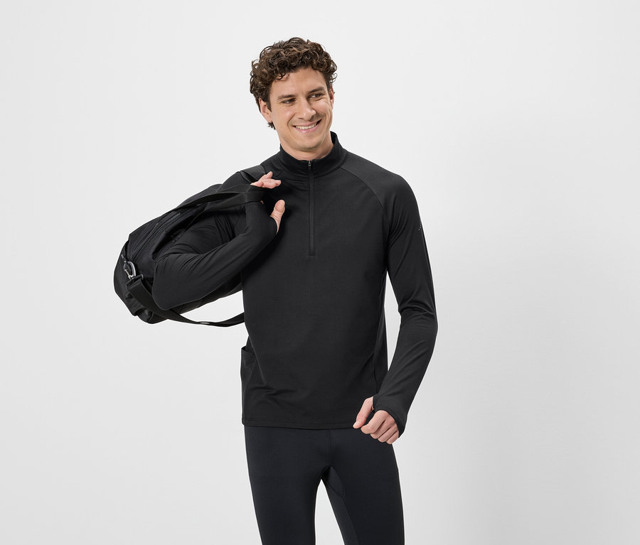 Un homme souriant aux cheveux bouclés, portant un haut de sport noir zippé et un pantalon noir, avec un sac de sport noir sur l'épaule.