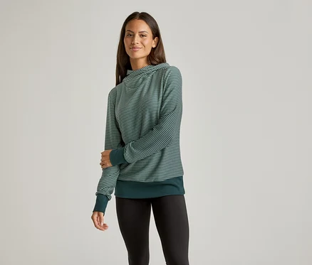 Une femme aux cheveux longs porte un pull rayé vert et un legging noir sur un fond neutre.