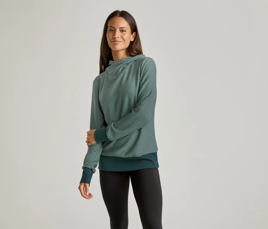 Une femme aux cheveux longs porte un pull rayé vert et un legging noir sur un fond neutre.
