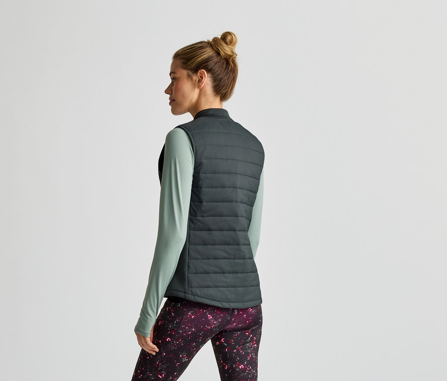 Femme vue de dos portant un gilet vert, un t-shirt et un legging à motifs sur fond clair.