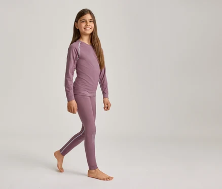 Une jeune fille souriante aux longs cheveux bruns marche pieds nus, vêtue d'un haut à manches longues violet et d'un legging assorti avec des rayures claires.