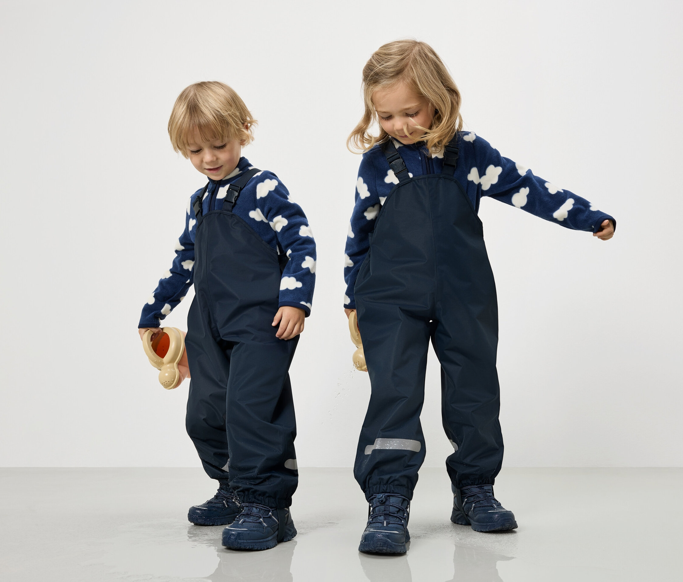 Deux enfants en salopettes bleu marine et pulls nuages bleu et blanc se tiennent sur un fond blanc.